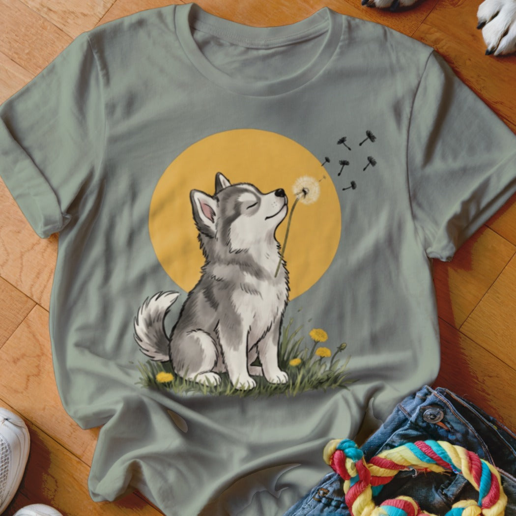 Sage green t-shirt featuring an illustration of a husky sitting in grass and blowing dandelion seeds.