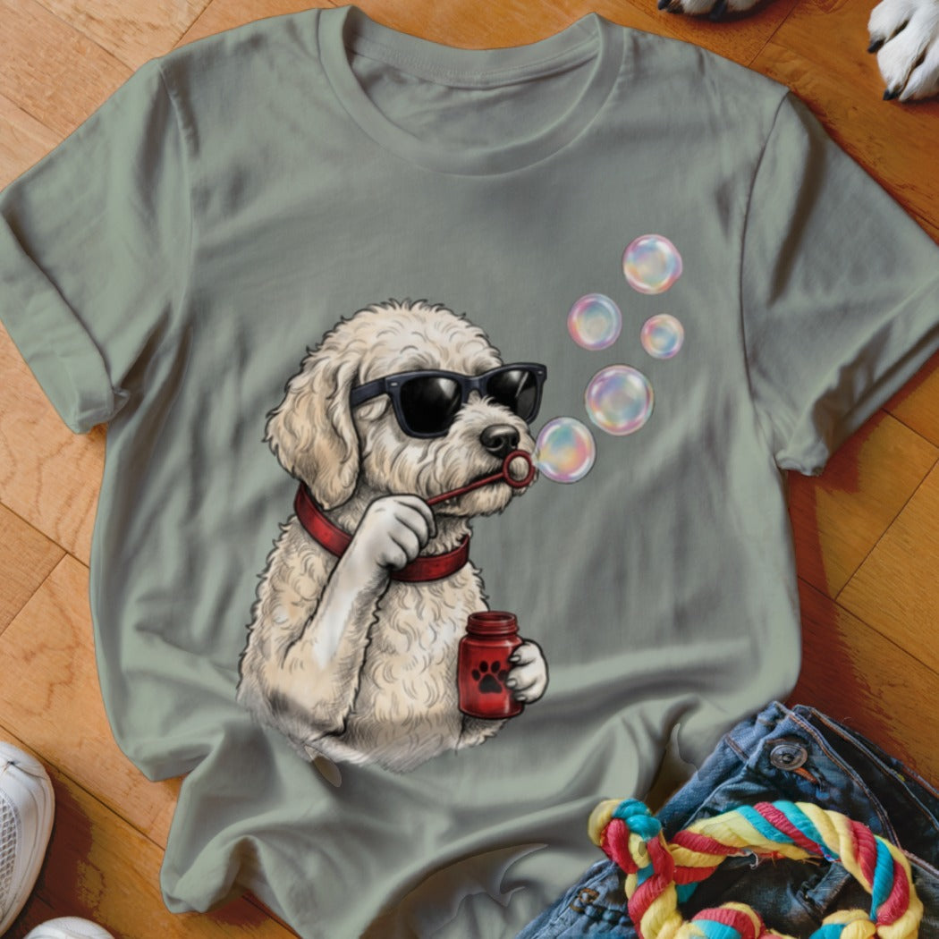 A sage green t-shirt featuring a graphic of a curly white dog wearing sunglasses and blowing bubbles.