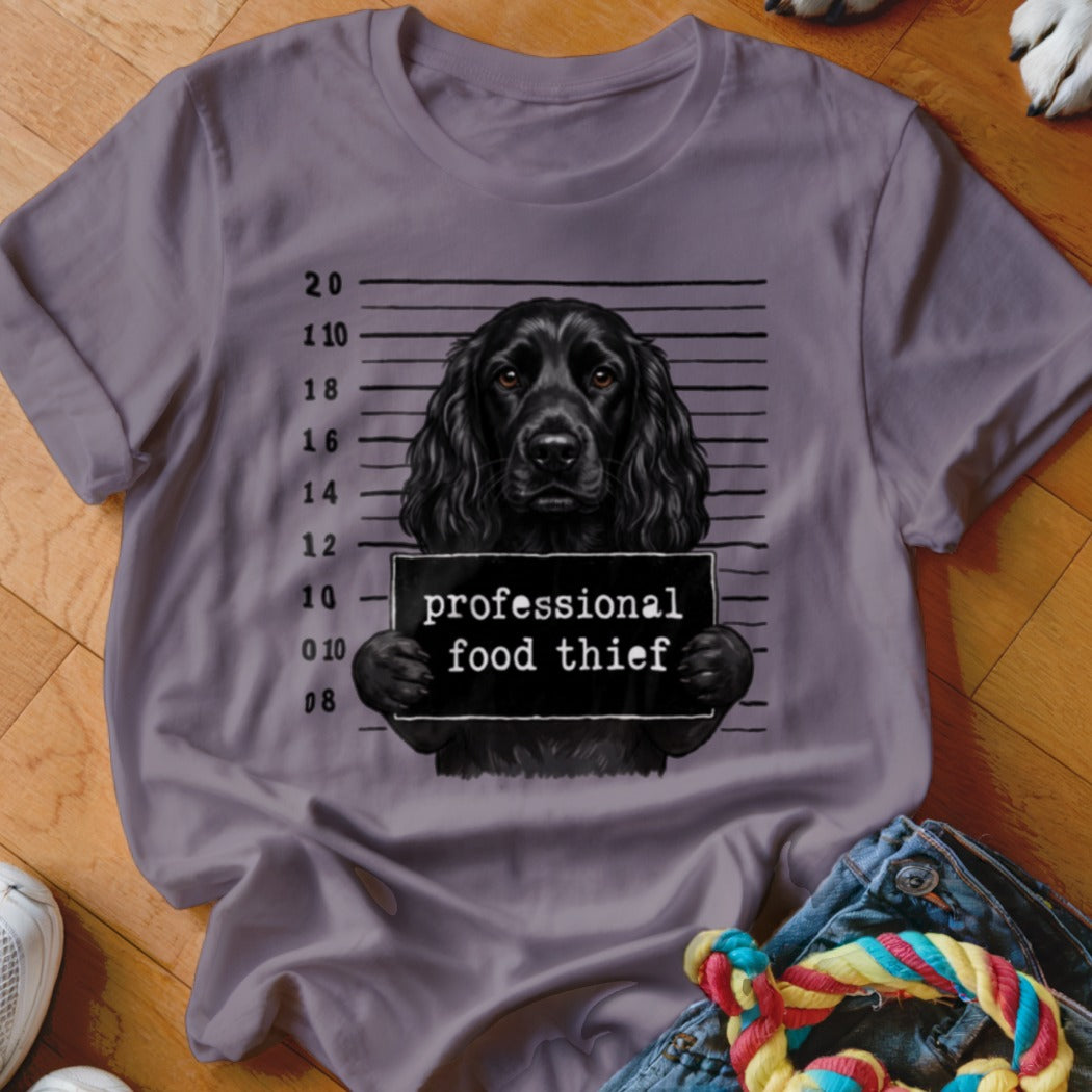 Black dog in mugshot photo holding a "professional food thief" sign on a purple t-shirt.