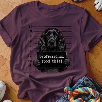 Black dog mugshot with "professional food thief" sign on a maroon t-shirt.
