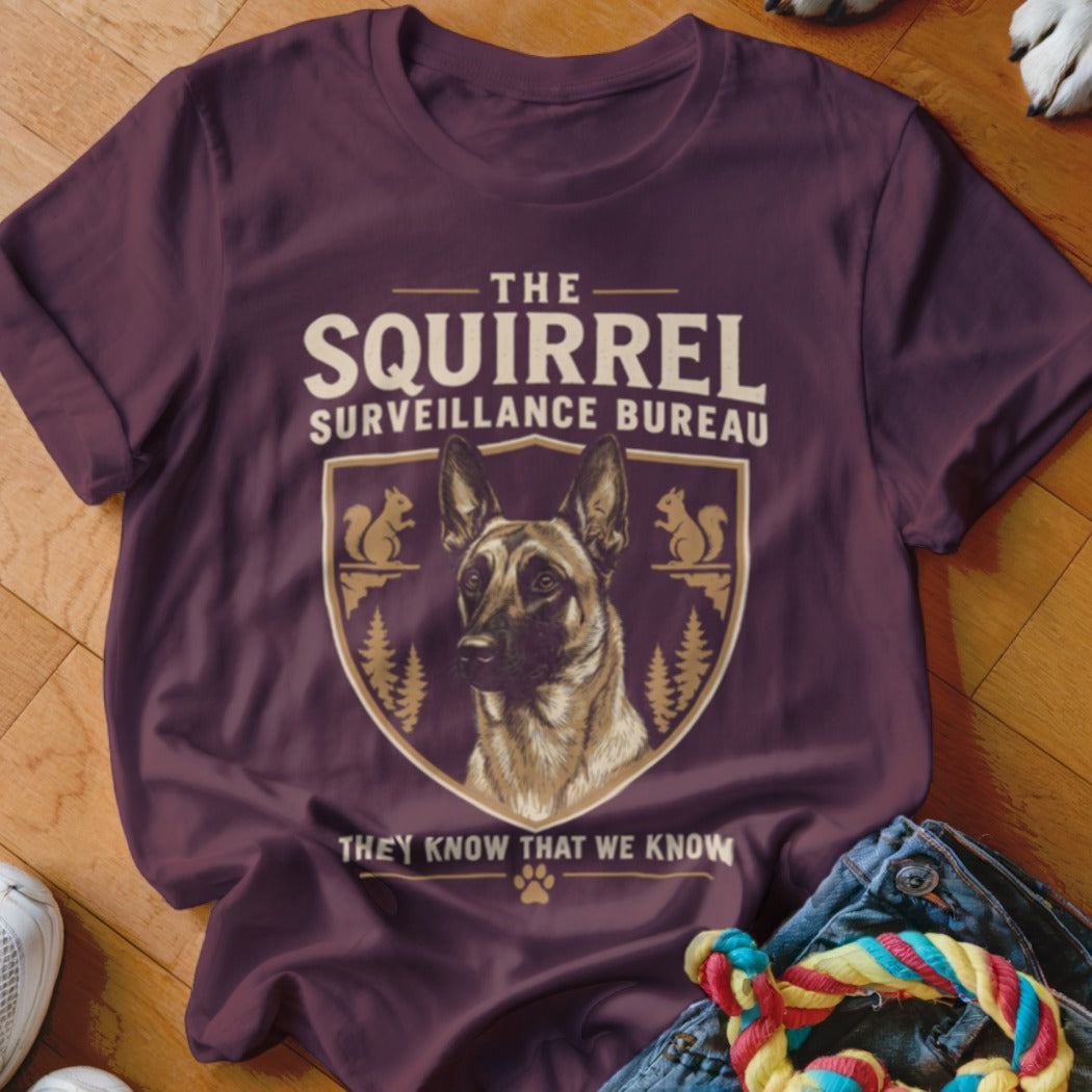 Maroon t-shirt featuring a crest with a German Shepherd, squirrels, pine trees, and text reading THE SQUIRREL SURVEILLANCE BUREAU THEY KNOW THAT WE KNOW.