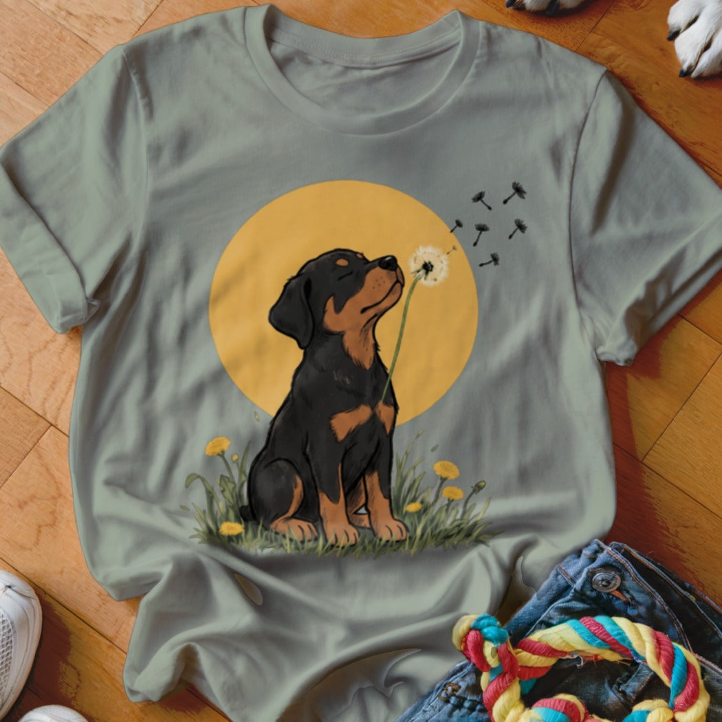 Sage green crewneck t-shirt featuring an illustrated rottweiler puppy blowing dandelion seeds against a yellow sun background.