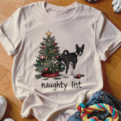 A light-colored t-shirt with a graphic of a black and white dog lifting its leg at a decorated Christmas tree, with the text naughty list underneath.