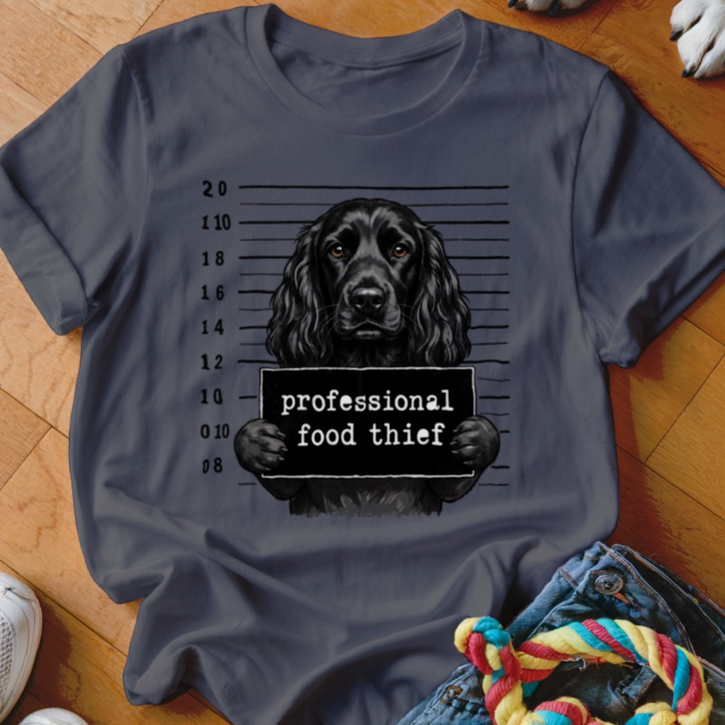 Black dog mugshot with "professional food thief" sign on t-shirt.