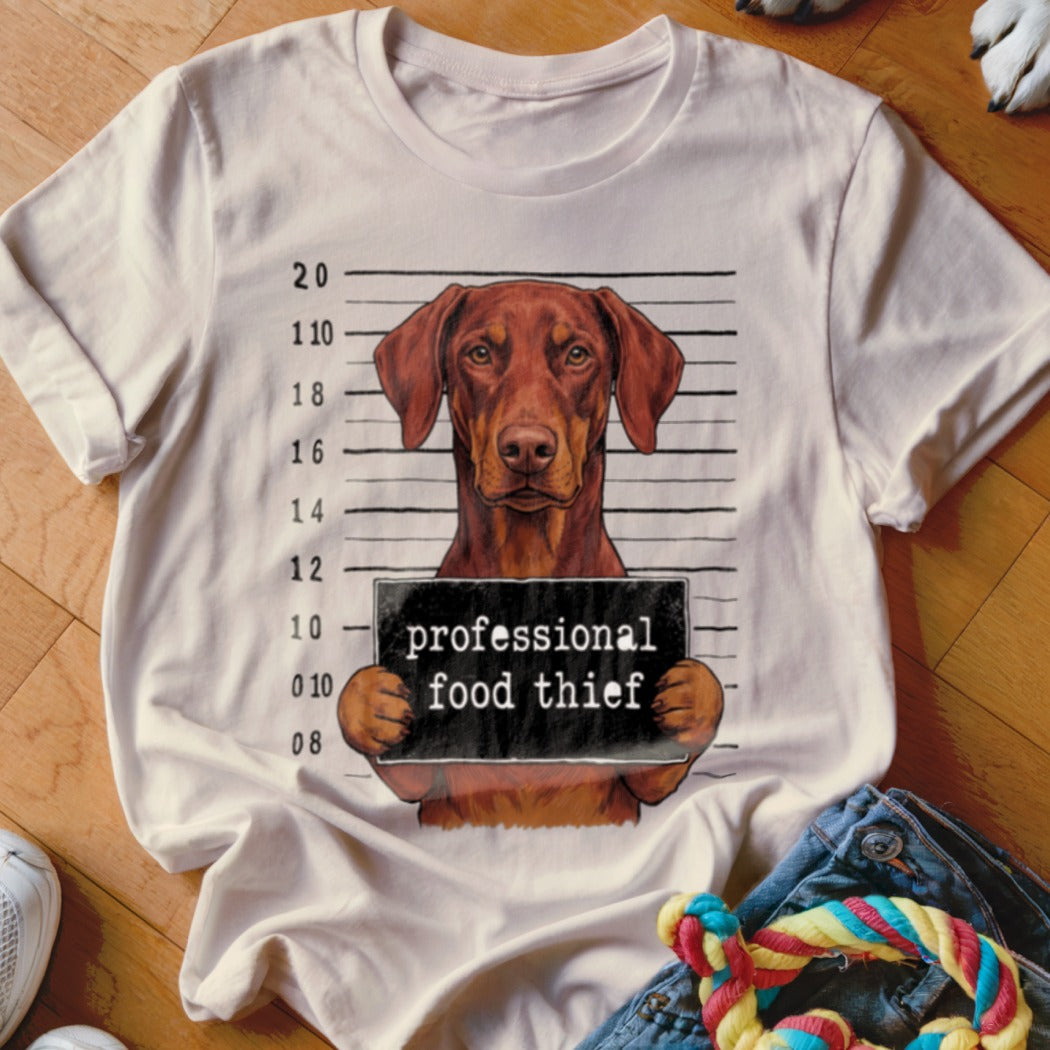 Brown dog mugshot with "professional food thief" sign on white t-shirt.