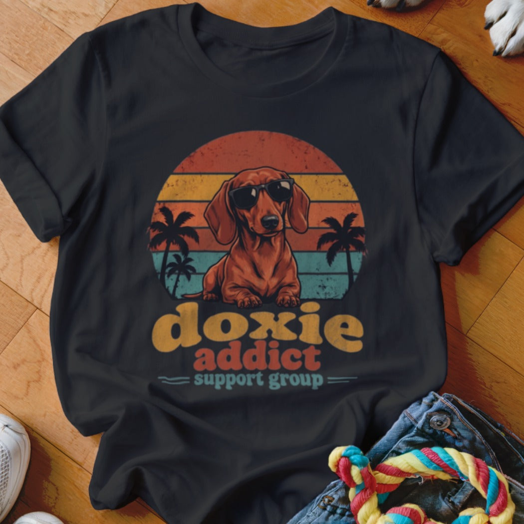 A black t-shirt features a graphic of a dachshund wearing sunglasses in front of a sunset, palm trees, and retro text reading doxie addict support group.