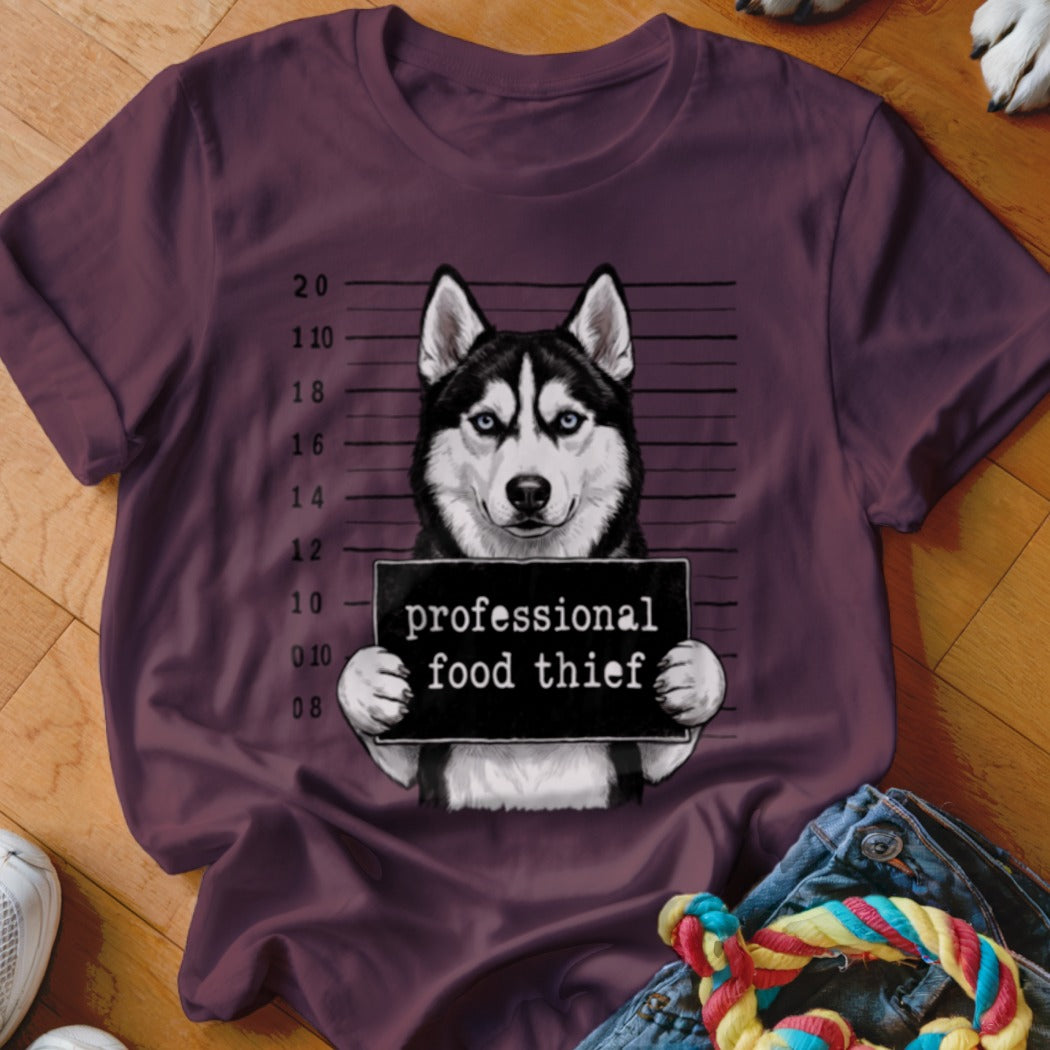 Husky holding a "professional food thief" sign in mugshot style on a dark red t-shirt.
