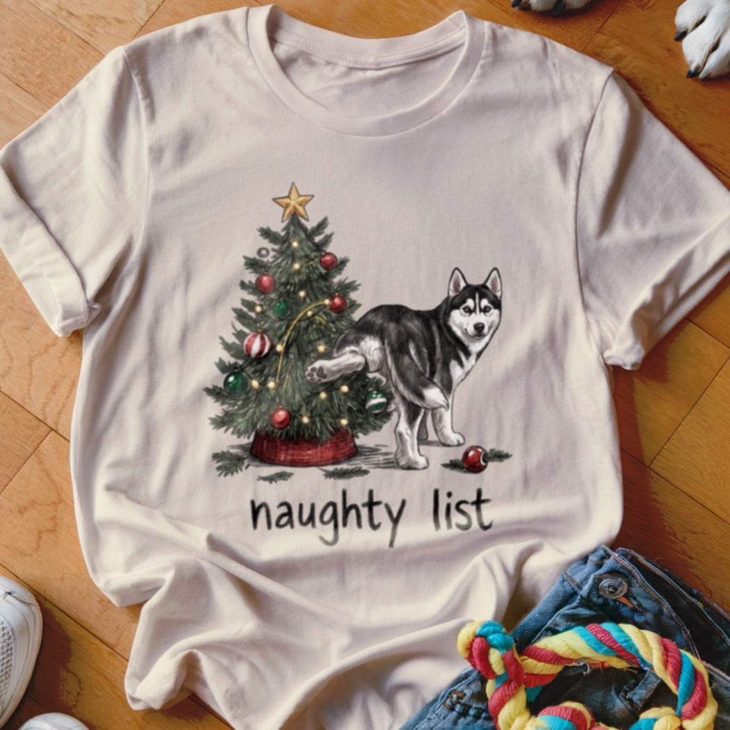 A cream-colored t-shirt featuring a cartoon illustration of a husky lifting its leg against a decorated Christmas tree above the text "naughty list".