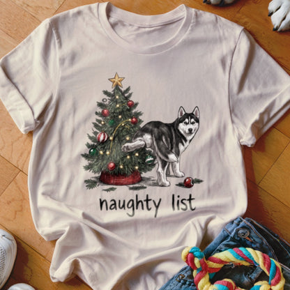 A cream-colored t-shirt featuring a cartoon illustration of a husky lifting its leg against a decorated Christmas tree above the text "naughty list".
