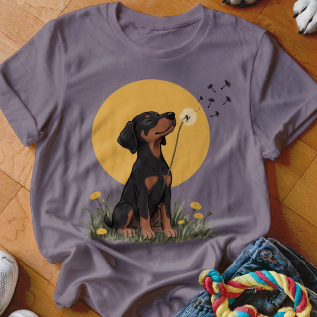 A purple short-sleeved t-shirt featuring an illustration of a black and tan puppy blowing dandelion seeds.
