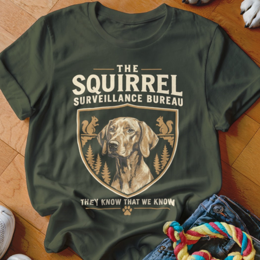 Dark green t-shirt featuring a dog graphic, pine trees, squirrels, and text reading The Squirrel Surveillance Bureau.