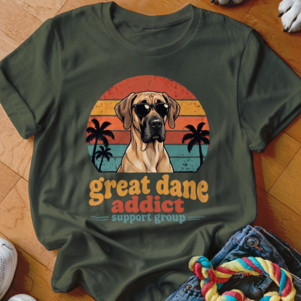 An olive green t-shirt displays a graphic of a Great Dane wearing sunglasses against a retro sunset and palm tree background, with the text Great Dane Addict Support Group.