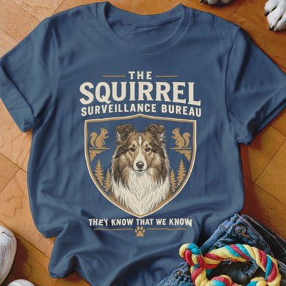 A blue t-shirt printed with a graphic featuring a Sheltie dog, squirrels, and the text The Squirrel Surveillance Bureau.