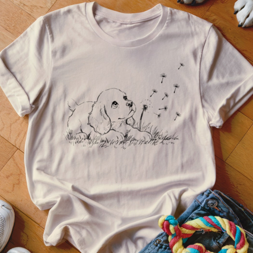 A light pink t-shirt featuring a black line drawing of a puppy watching dandelion seeds blow away.