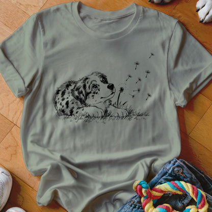 Sage green t-shirt featuring a black line art graphic of a spotted puppy looking at dandelion seeds.