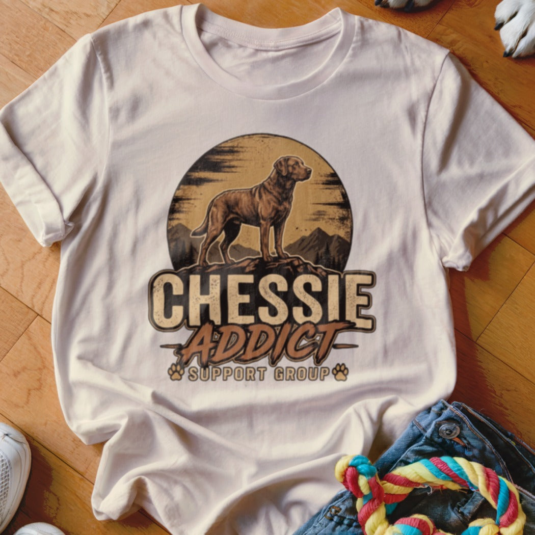 A light cream t-shirt featuring a vintage-style graphic of a Chesapeake Bay Retriever standing on a mountain peak, with the text Chessie Addict Support Group.
