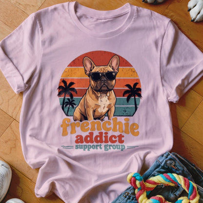 A light pink t-shirt featuring a graphic of a French bulldog wearing sunglasses before a sunset and palm tree background, with text reading "frenchie addict support group."