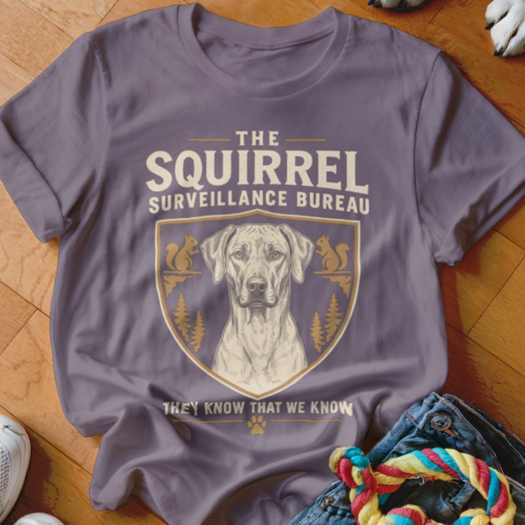 A purple t-shirt featuring a dog graphic inside a crest with squirrels and trees, and the text The Squirrel Surveillance Bureau.