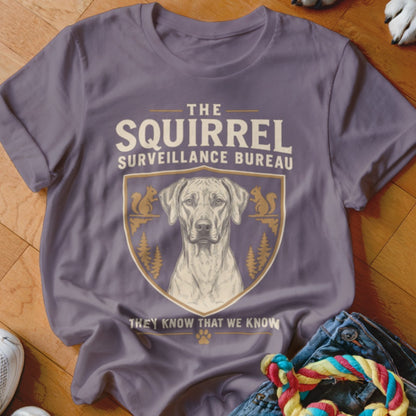 A purple t-shirt featuring a dog graphic inside a crest with squirrels and trees, and the text The Squirrel Surveillance Bureau.
