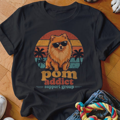A black t-shirt printed with a Pomeranian wearing sunglasses in front of a retro sunset and palm trees, reading "pom addict support group".