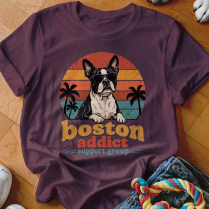 Maroon t-shirt featuring a retro graphic of a Boston Terrier wearing sunglasses in front of a sunset, with palm trees and the text boston addict support group.