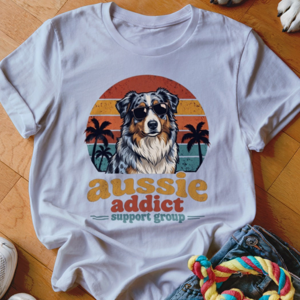 A white t-shirt featuring an Australian Shepherd wearing sunglasses in front of a retro sunset graphic.
