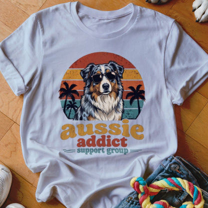 A white t-shirt featuring an Australian Shepherd wearing sunglasses in front of a retro sunset graphic.