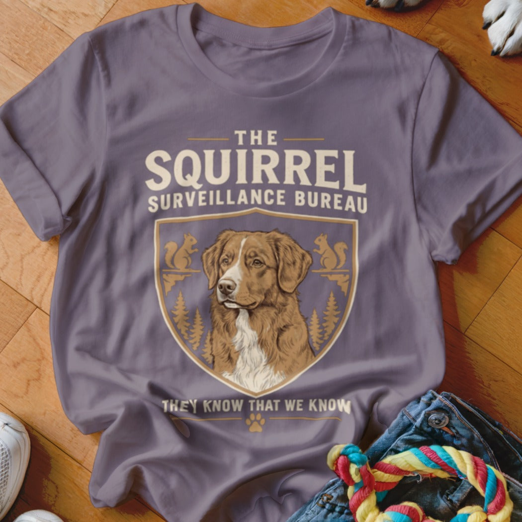 A purple t-shirt featuring a graphic with a dog, squirrels, trees, and the text "The Squirrel Surveillance Bureau".