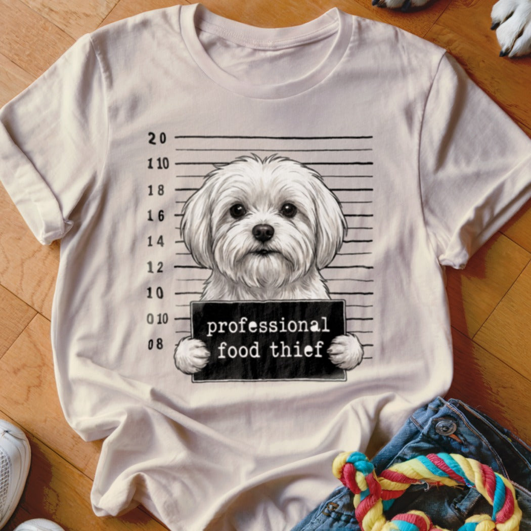 White Maltese dog mugshot with "professional food thief" sign on a t-shirt.