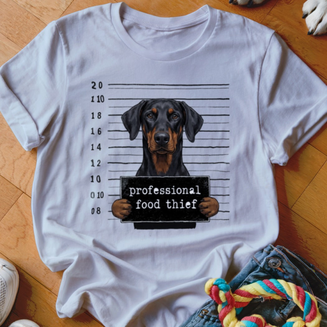 Doberman dog mugshot with "professional food thief" sign on white t-shirt.
