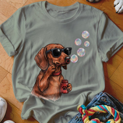 Sage green t-shirt featuring a graphic of a dachshund wearing sunglasses and blowing soap bubbles.