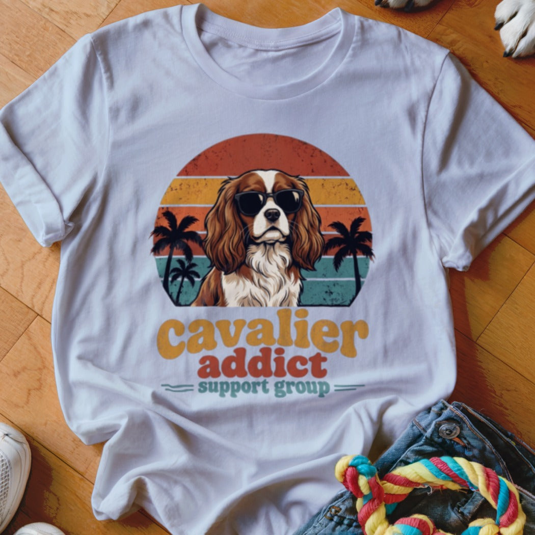 White t-shirt featuring a retro graphic of a Cavalier King Charles Spaniel wearing sunglasses with palm trees and sunset stripes.