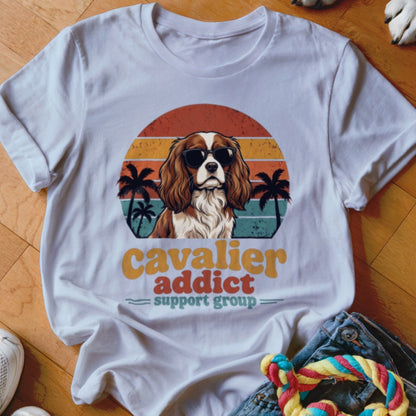 White t-shirt featuring a retro graphic of a Cavalier King Charles Spaniel wearing sunglasses with palm trees and sunset stripes.