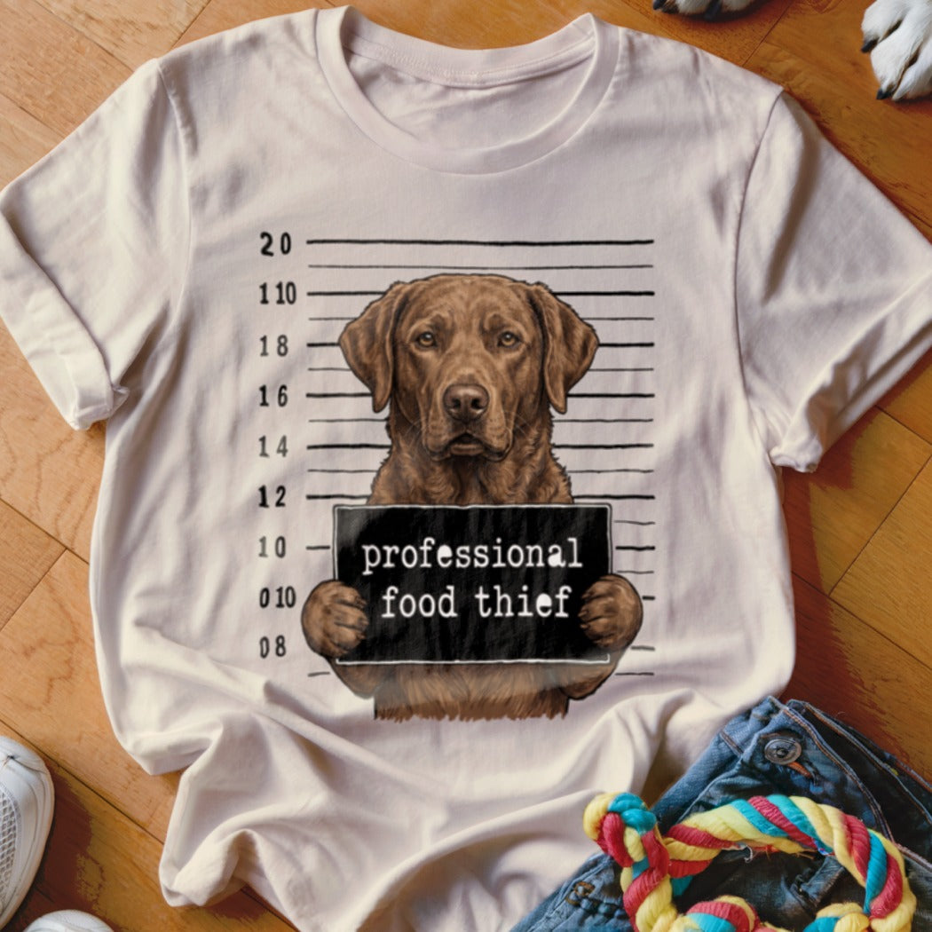 Brown lab dog mugshot with "professional food thief" sign on t-shirt