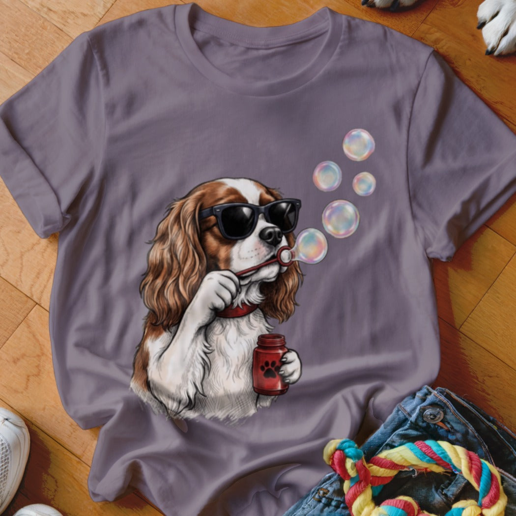 A purple t-shirt featuring a graphic of a Cavalier King Charles Spaniel wearing sunglasses and blowing bubbles.