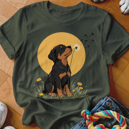 An olive green t-shirt featuring an illustration of a Rottweiler puppy blowing a dandelion against a yellow circle.