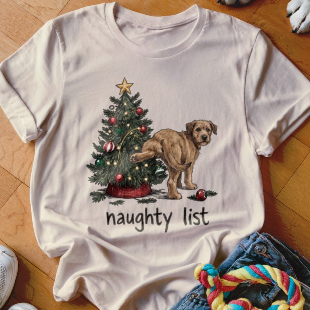Beige short-sleeve t-shirt with a graphic of a dog lifting its leg at a decorated Christmas tree and the text naughty list.