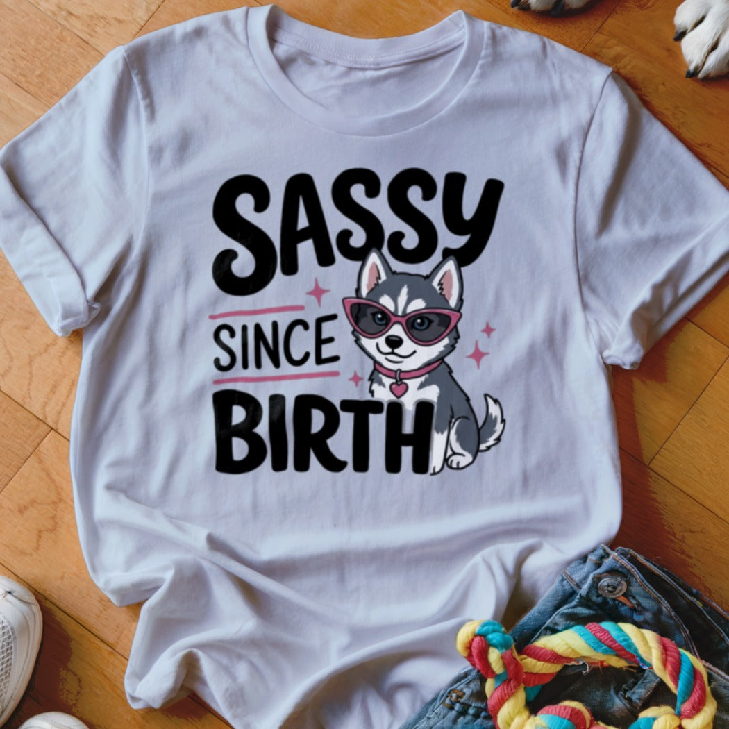 White t-shirt with graphic of a husky puppy wearing sunglasses and text "SASSY SINCE BIRTH".