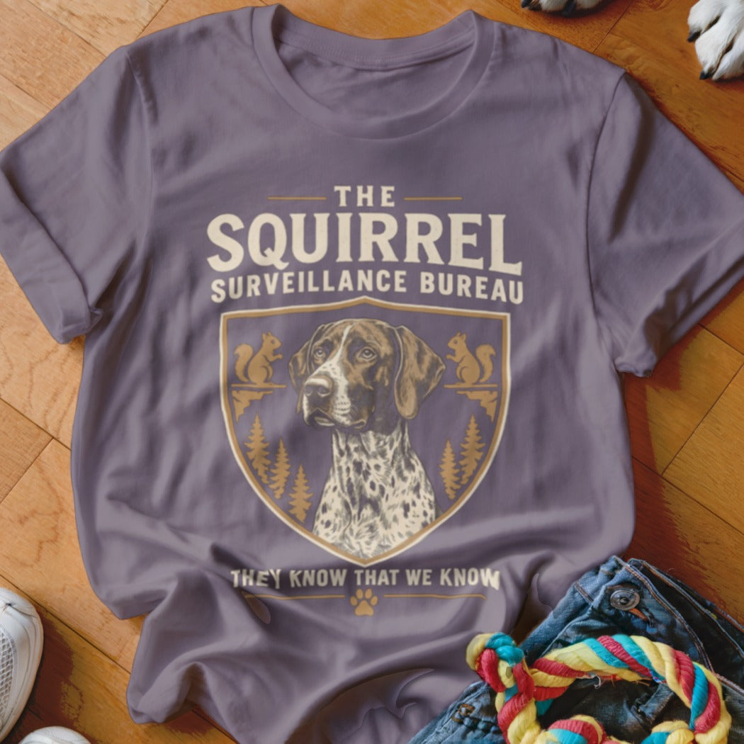 A purple t-shirt featuring a graphic with a pointing dog, squirrels, and the text The Squirrel Surveillance Bureau.