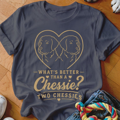 A dark gray t-shirt featuring a golden outline of two Chesapeake Bay Retrievers inside a heart and the text "What's better than a Chessie? Two Chessies."