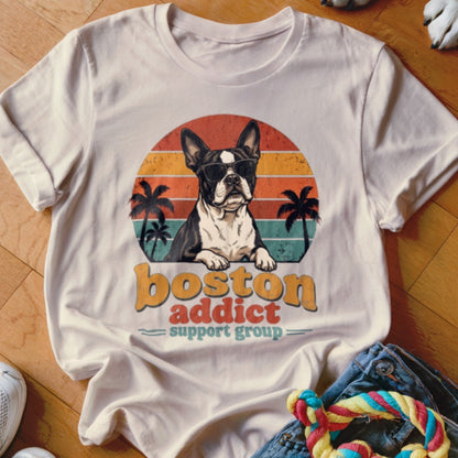 Beige t-shirt featuring a retro graphic of a sunglasses-wearing Boston terrier with palm trees and text reading boston addict support group.