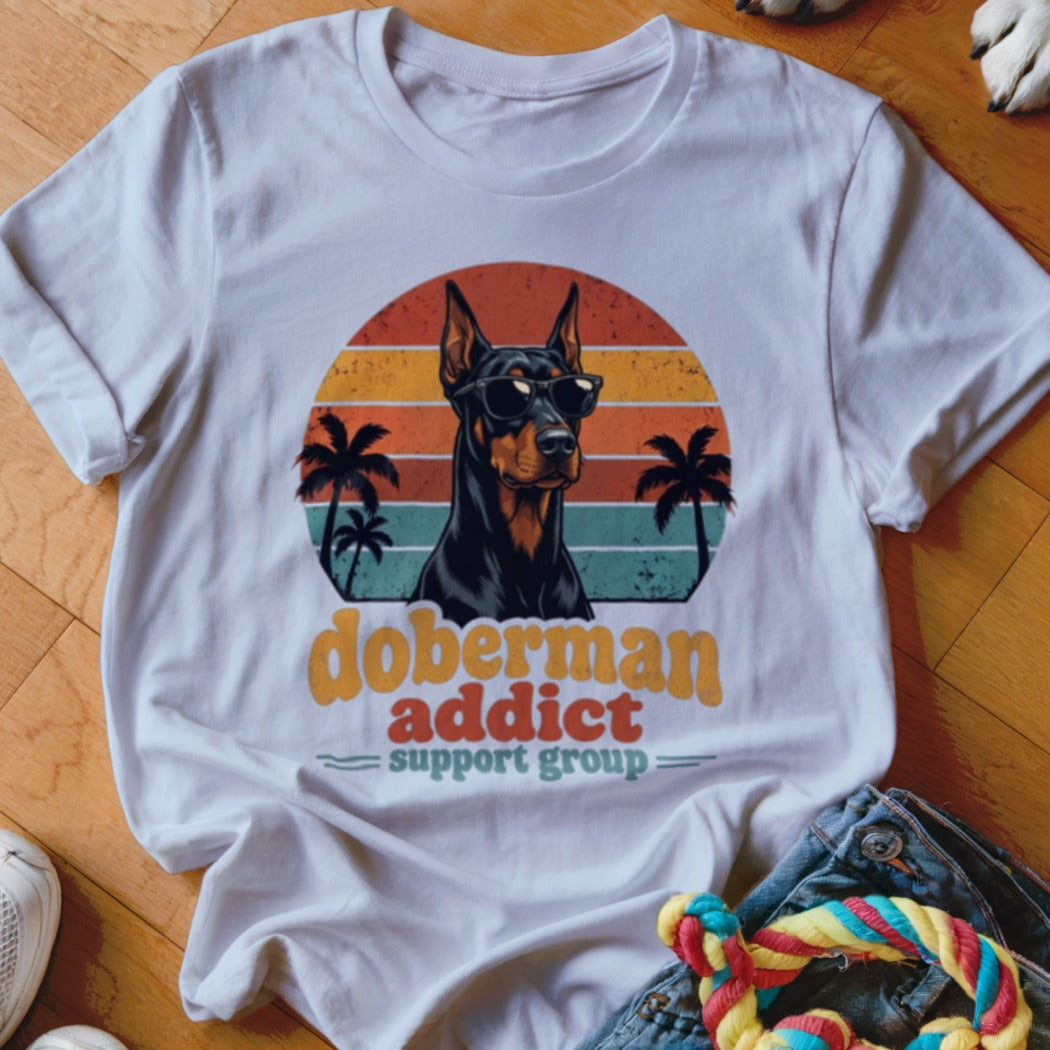 A white t-shirt featuring a retro sunset graphic, a Doberman wearing sunglasses, and text reading "Doberman Addict Support Group."