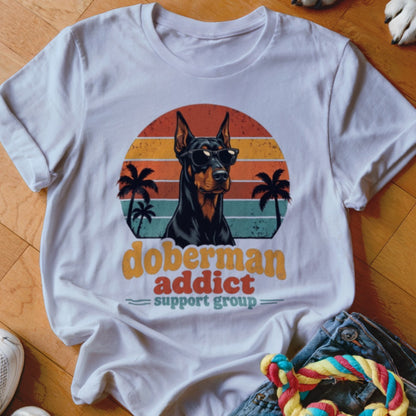 A white t-shirt featuring a retro sunset graphic, a Doberman wearing sunglasses, and text reading "Doberman Addict Support Group."
