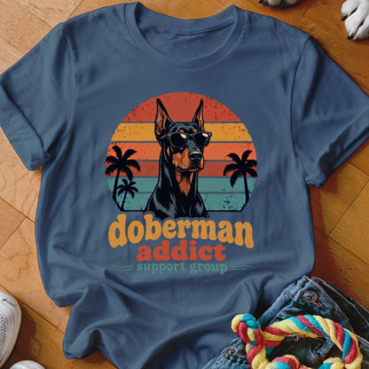 A slate blue t-shirt featuring a retro graphic of a Doberman wearing sunglasses before palm trees and a sunset.