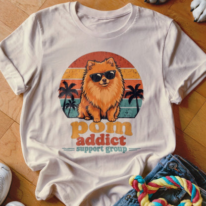 A beige t-shirt with a graphic of a Pomeranian wearing sunglasses, palm trees, and text reading Pom Addict Support Group.