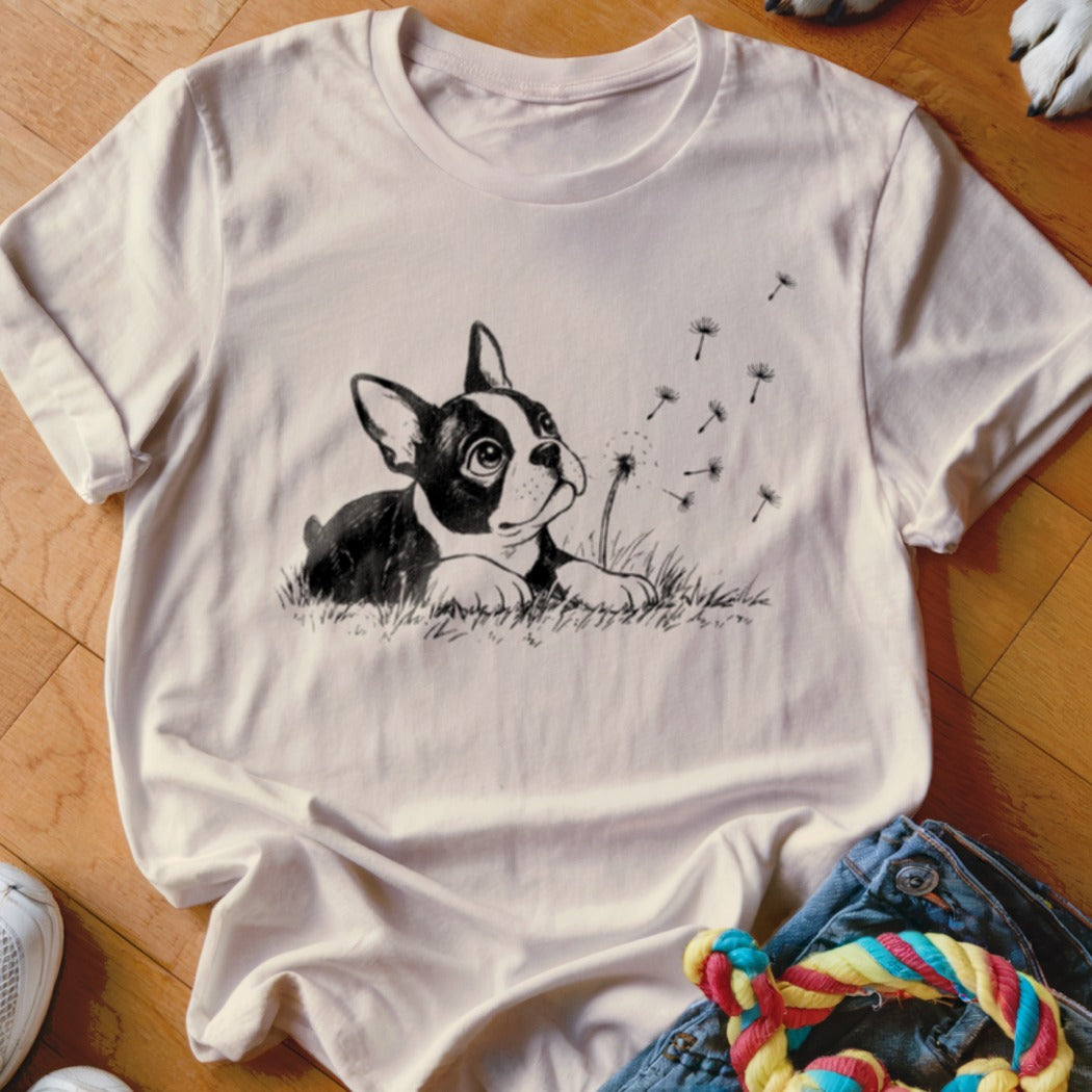 Light pink short-sleeved t-shirt featuring a black and white illustration of a dog in grass blowing dandelion seeds.