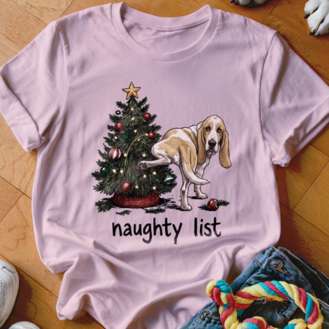 Basset hound near Christmas tree with text "naughty list" on a pink t-shirt.