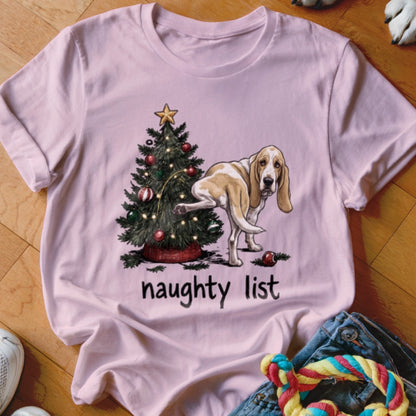 Basset hound near Christmas tree with text "naughty list" on a pink t-shirt.