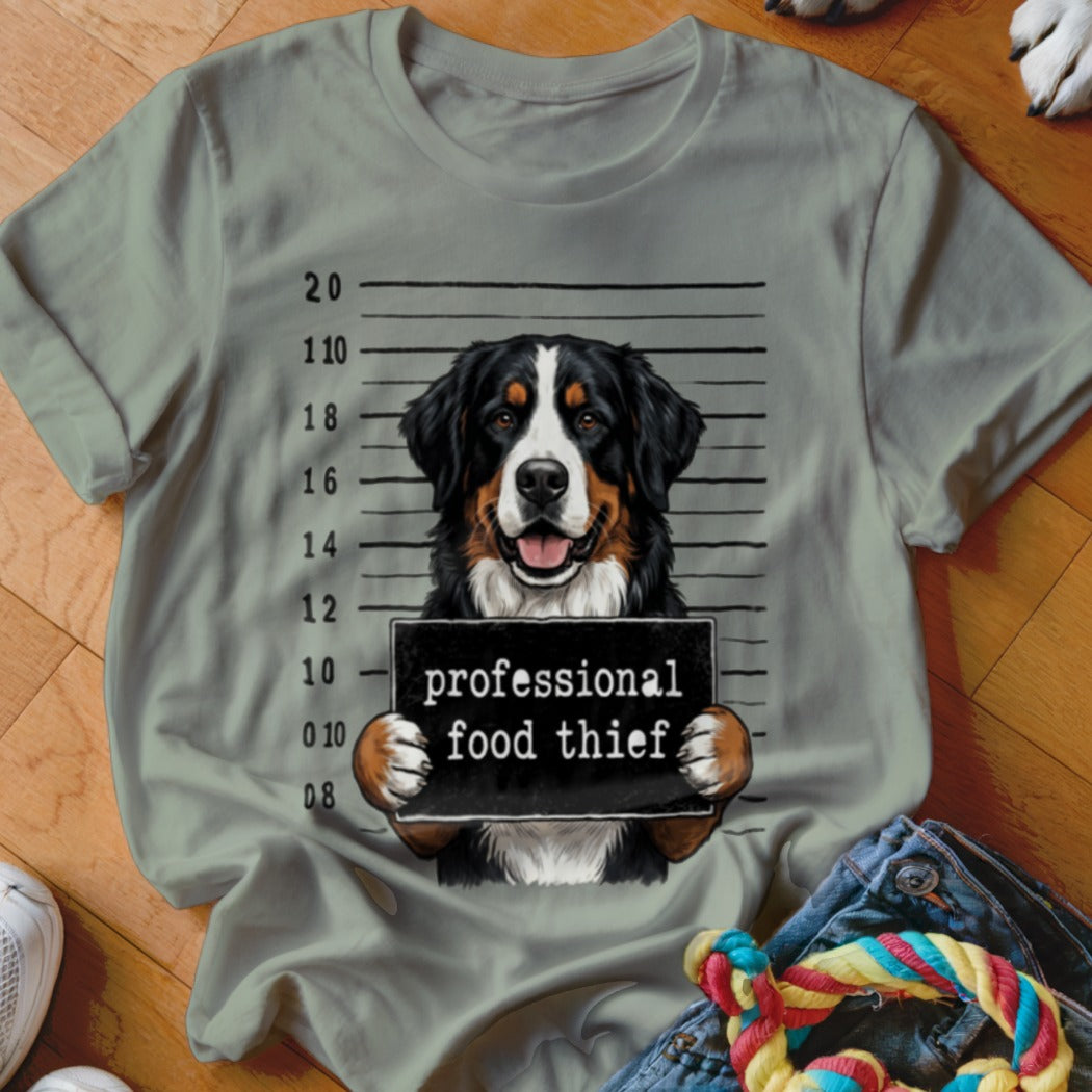 Bernese Mountain Dog mugshot holding "professional food thief" sign on a grey t-shirt.