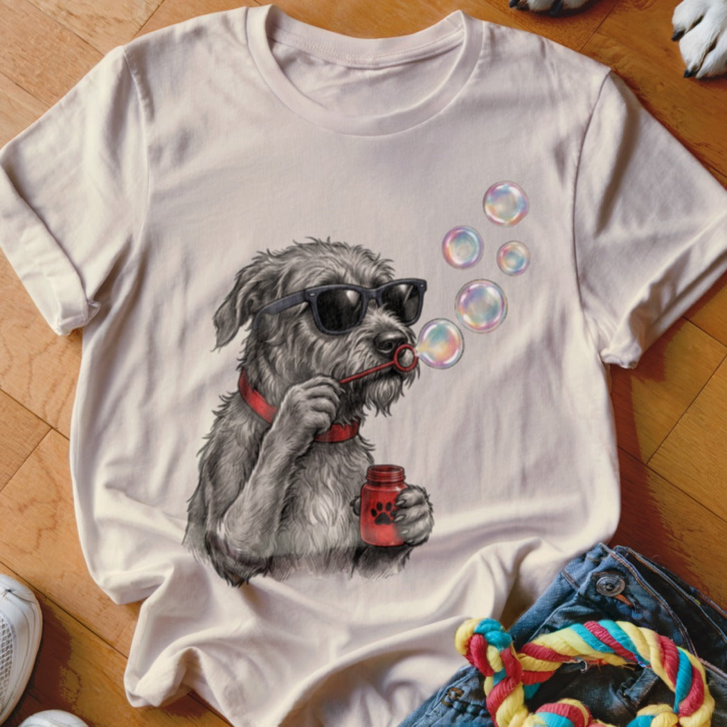 A beige t-shirt lies on a wooden floor, featuring a graphic of a scruffy dog wearing sunglasses and blowing bubbles.